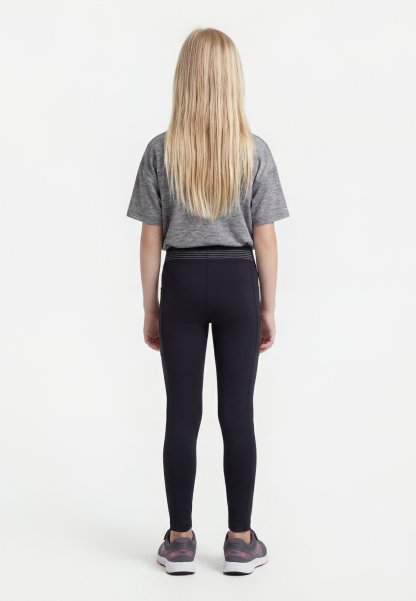 Niña rubia de pie de espaldas, con camiseta gris, mallas negras y zapatillas grises con detalles en rosa sobre un fondo blanco.