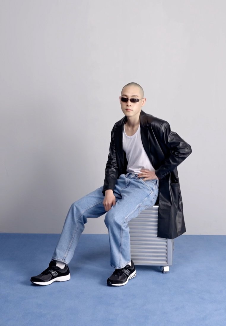 Chaqueta de cuero negra, camiseta blanca sin mangas, jeans de mezclilla azul claro, zapatillas negras. Sentado sobre un estuche metálico con un fondo sencillo.
