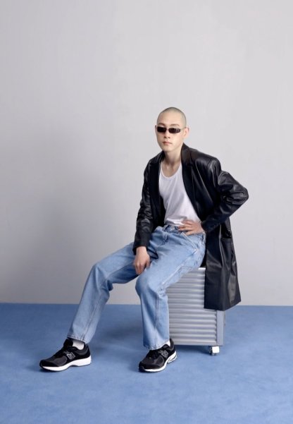 Chaqueta de cuero negra, camiseta blanca sin mangas, jeans de mezclilla azul claro, zapatillas negras. Sentado sobre un estuche metálico con un fondo sencillo.