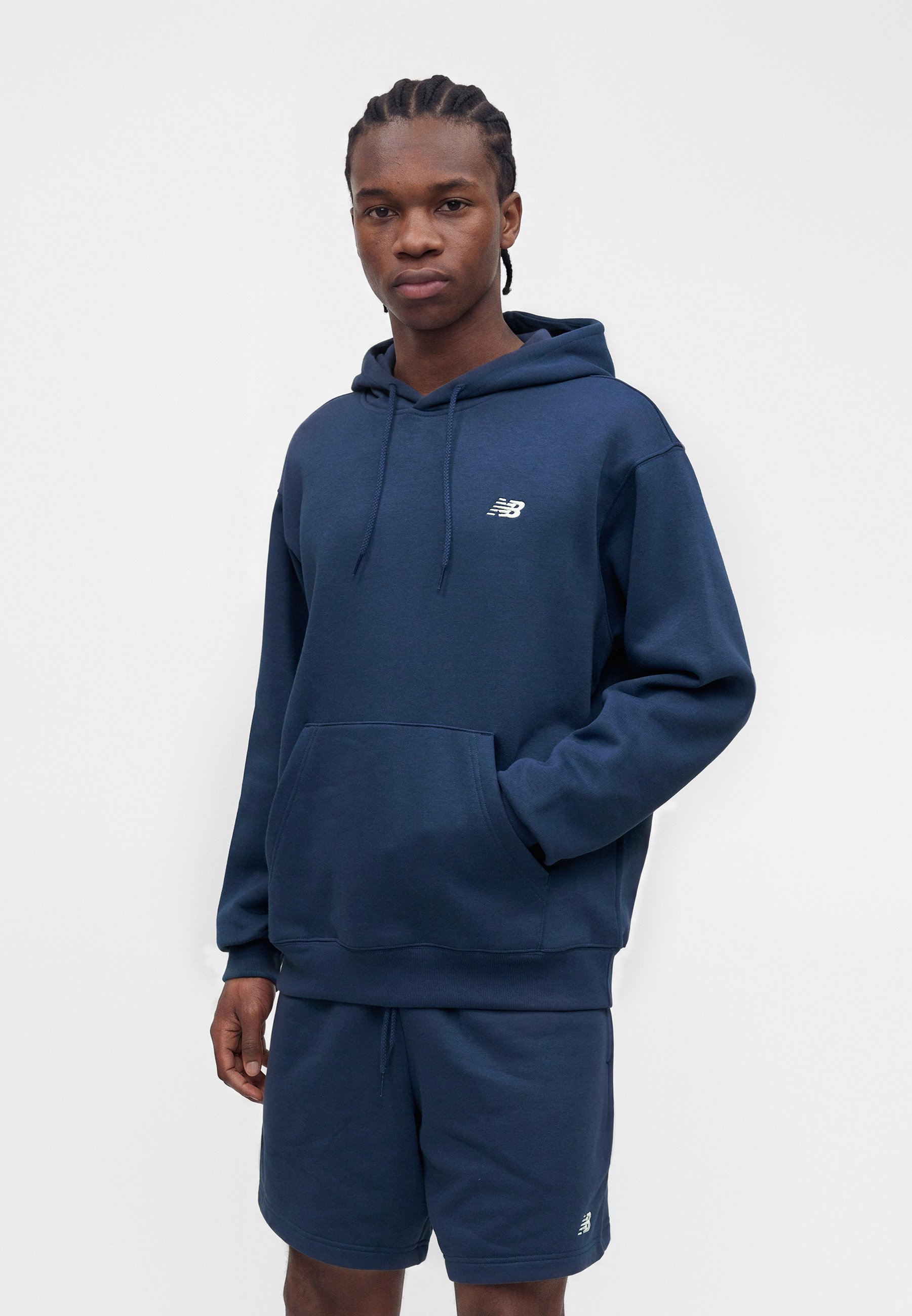 Joven con el pelo trenzado que lleva una sudadera con capucha azul marino de New Balance y pantalones cortos a juego sobre un fondo blanco liso.