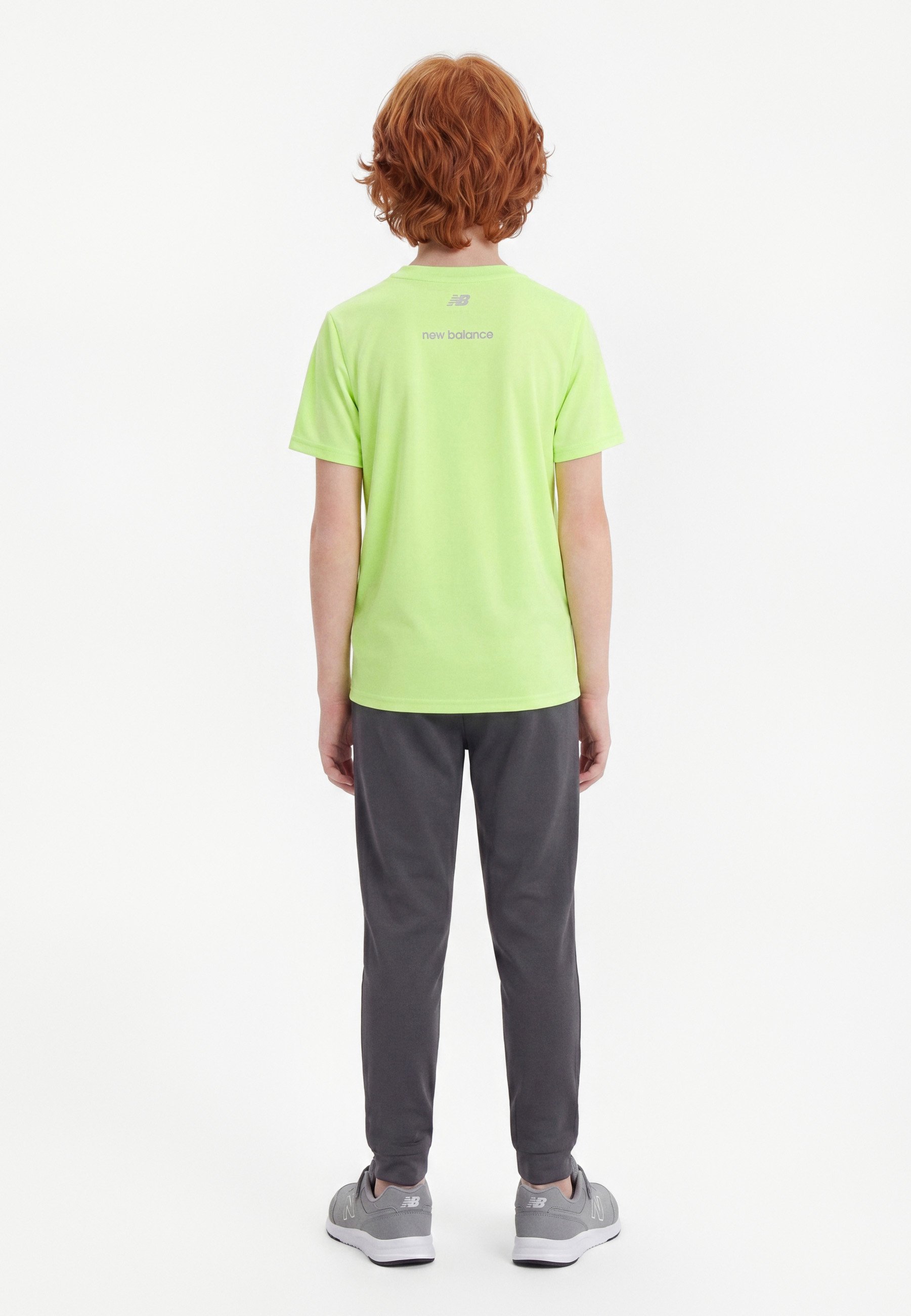 Niño con cabello rojo que lleva una camiseta lime verde de New Balance, pantalones gris oscuro y zapatillas grises, de pie de espaldas contra un fondo liso.