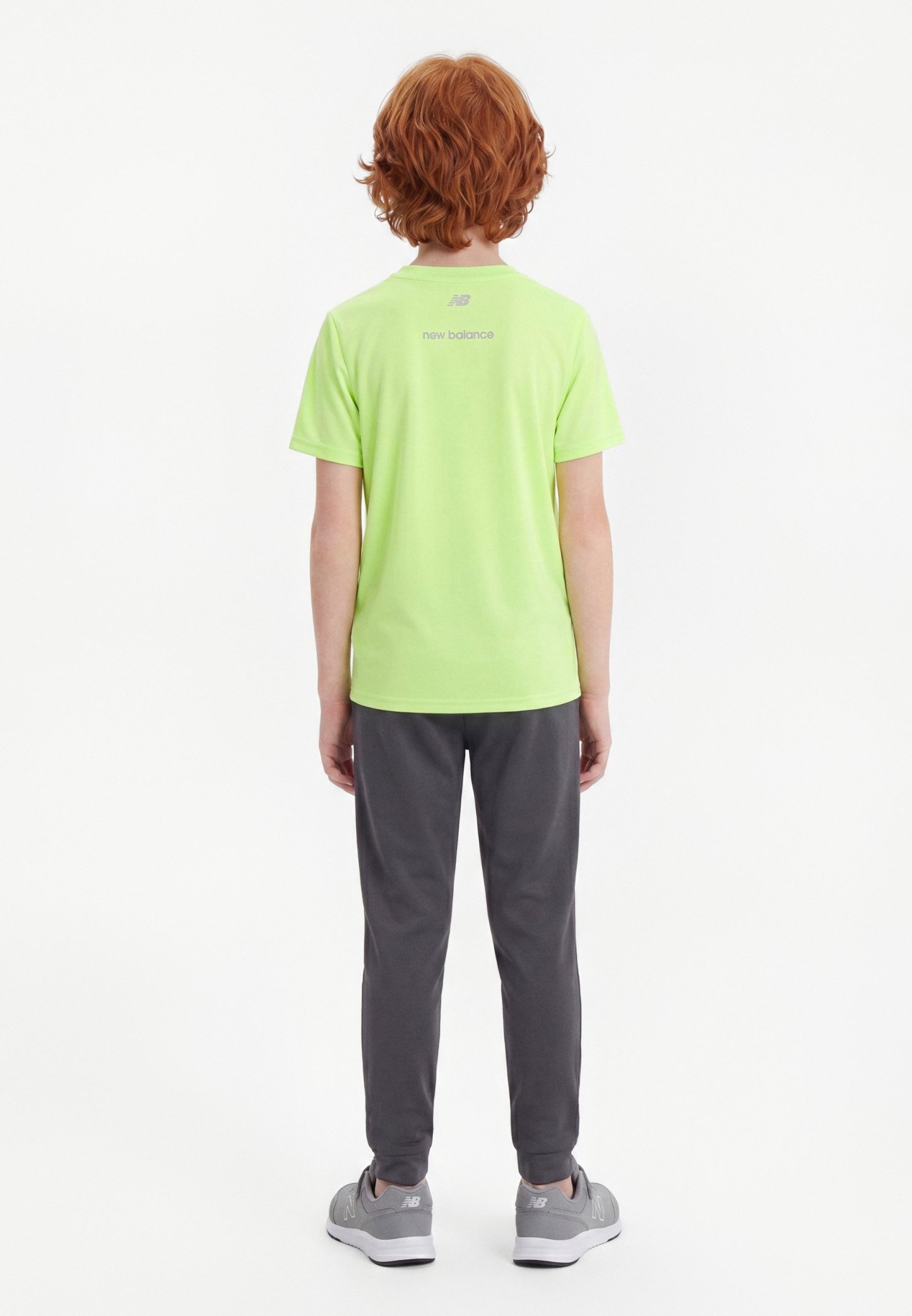 Niño con cabello rojo que lleva una camiseta lime verde de New Balance, pantalones gris oscuro y zapatillas grises, de pie de espaldas contra un fondo liso.