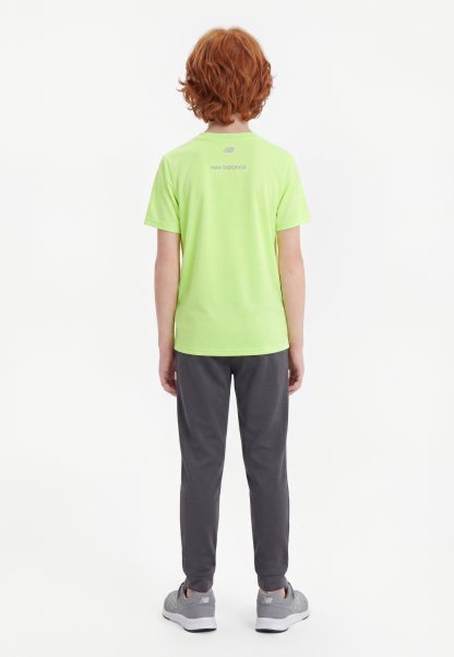 Niño con cabello rojo que lleva una camiseta lime verde de New Balance, pantalones gris oscuro y zapatillas grises, de pie de espaldas contra un fondo liso.