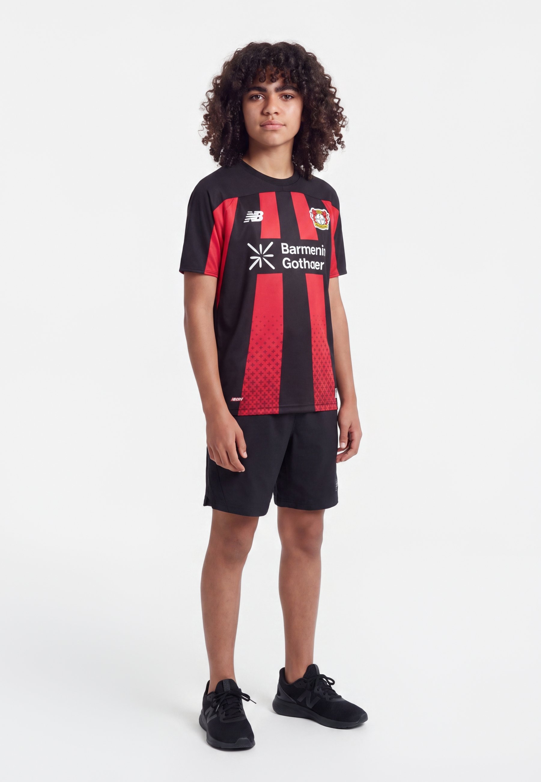 Joven con pelo rizado que lleva una camiseta de fútbol del Bayer Leverkusen negra y roja, pantalones cortos negros y zapatillas negras, de pie sobre un fondo blanco.
