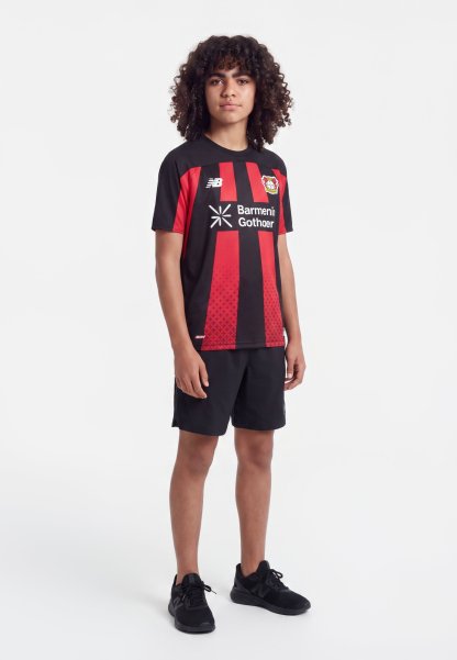 Joven con pelo rizado que lleva una camiseta de fútbol del Bayer Leverkusen negra y roja, pantalones cortos negros y zapatillas negras, de pie sobre un fondo blanco.