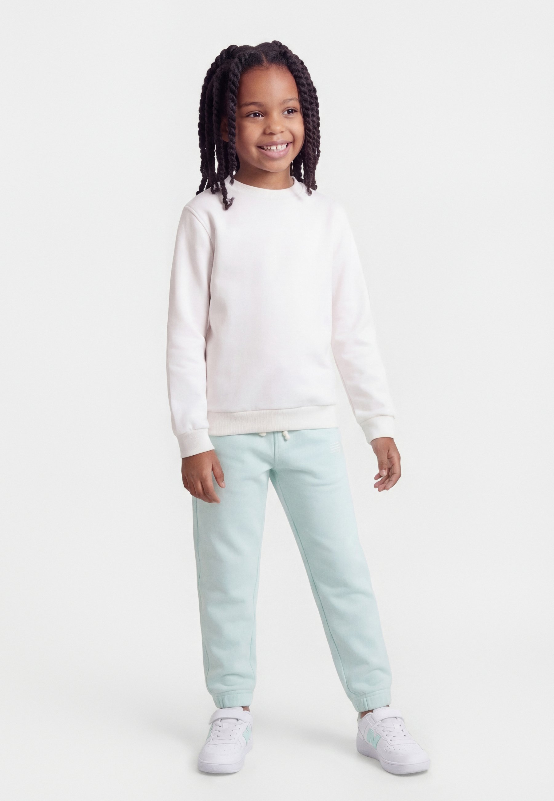 Niño sonriente con el cabello trenzado, vistiendo sudadera blanca, pantalones deportivos azul claro y zapatillas blancas, de pie sobre un fondo blanco.