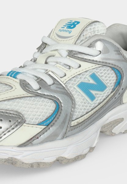 Zapatilla de running blanca y plateada de New Balance con detalles en azul claro, cordones atados y parte superior de malla texturizada.