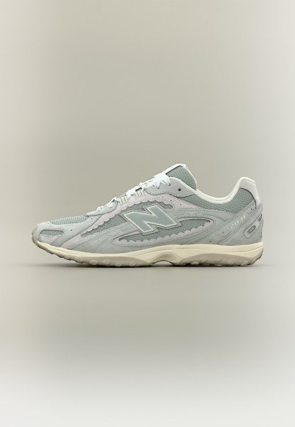 Zapatilla New Balance 204L en gris claro con parte superior de malla y ante, cordones blancos y suela texturizada beige sobre un fondo neutro y liso.