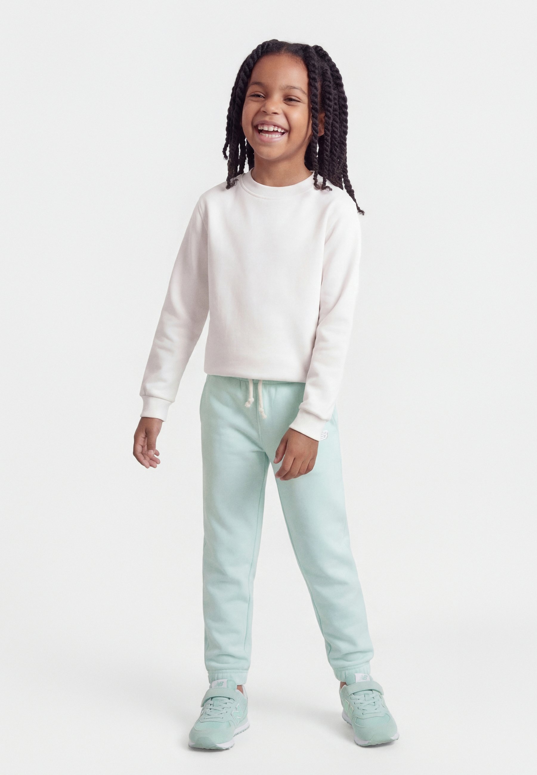 Niña sonriente con cabello largo trenzado, vestida con una sudadera blanca, pantalones deportivos azul claro y zapatillas a juego, posando frente a un fondo blanco liso.