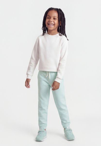 Niña sonriente con cabello largo trenzado, vestida con una sudadera blanca, pantalones deportivos azul claro y zapatillas a juego, posando frente a un fondo blanco liso.
