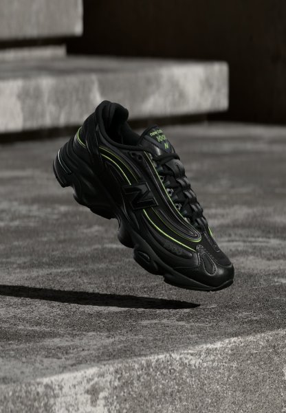 Zapatilla negra de New Balance con detalles en verde neón suspendida sobre escalones de hormigón gris con poca luz.