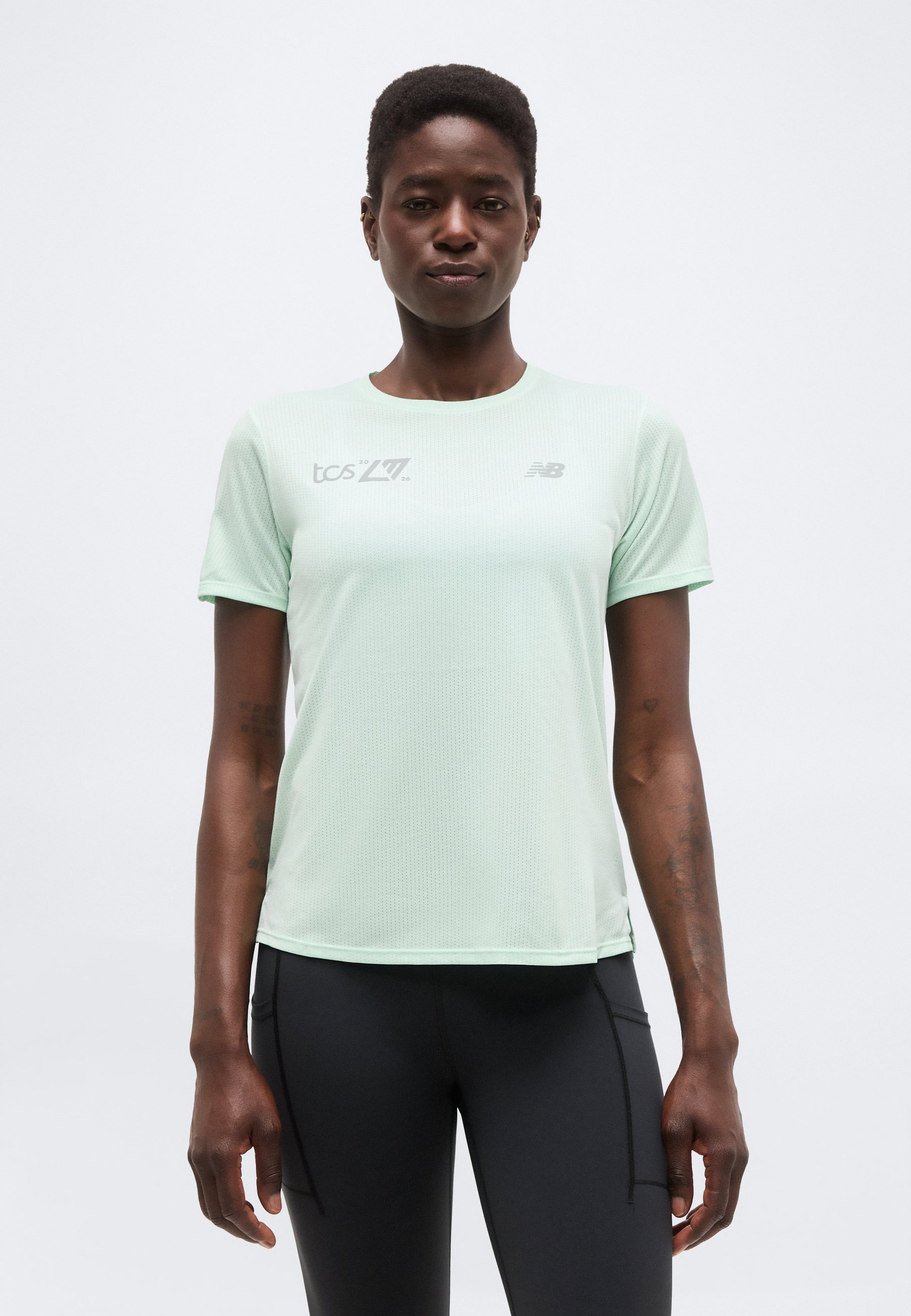 Mujer con una camiseta deportiva de manga corta color verde claro con logotipos y leggings negros, posando frente a un fondo blanco liso.