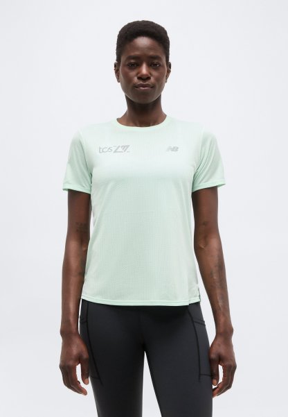 Mujer con una camiseta deportiva de manga corta color verde claro con logotipos y leggings negros, posando frente a un fondo blanco liso.