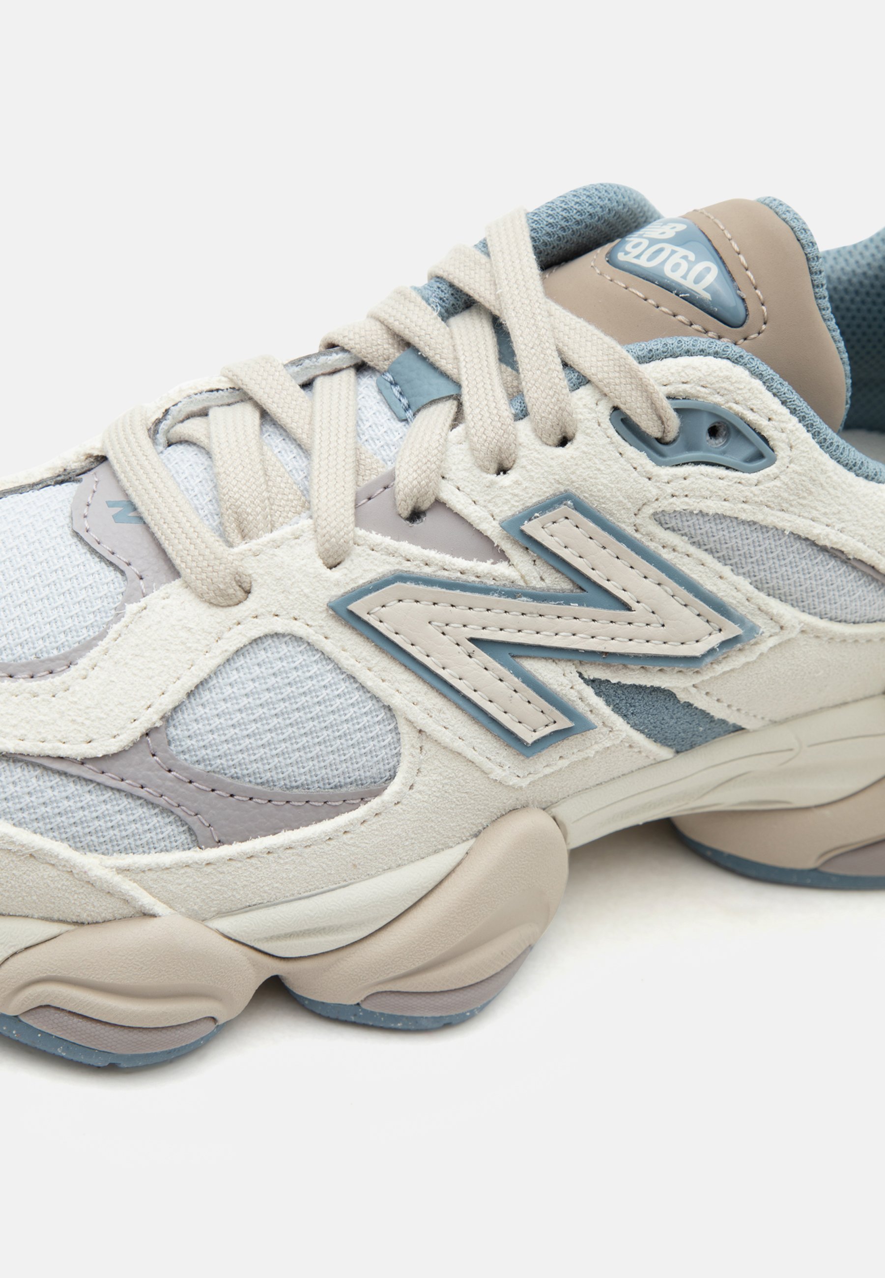 Zapatilla New Balance 9060 en beige, gris y azul con parte superior de malla y ante, suela acolchada y cordones beige atados en un lazo.
