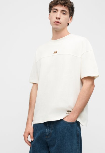 Joven con camisa blanca de manga corta y suelta con logo y vaqueros azules, de pie con una mano en el bolsillo frente a un fondo liso.