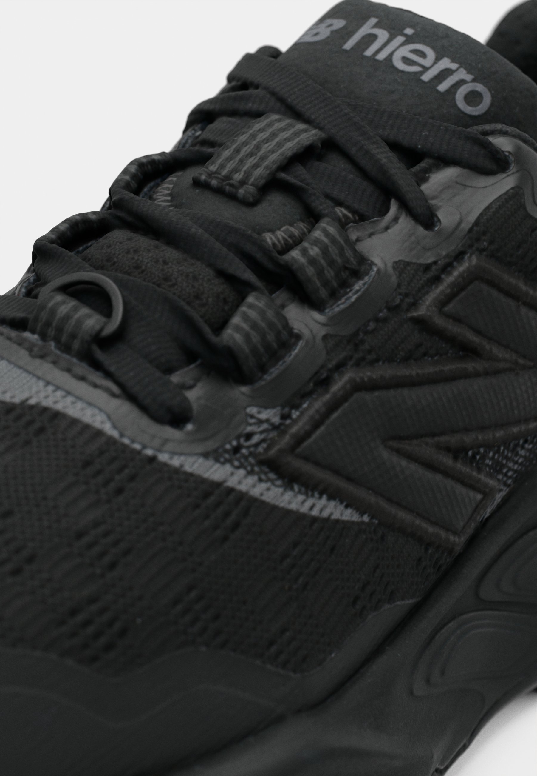 Zapatilla deportiva negra con parte superior texturizada, acentos lisos y un sistema de cordones pronunciado. Logotipo ubicado en la lengüeta, diseño elegante.
