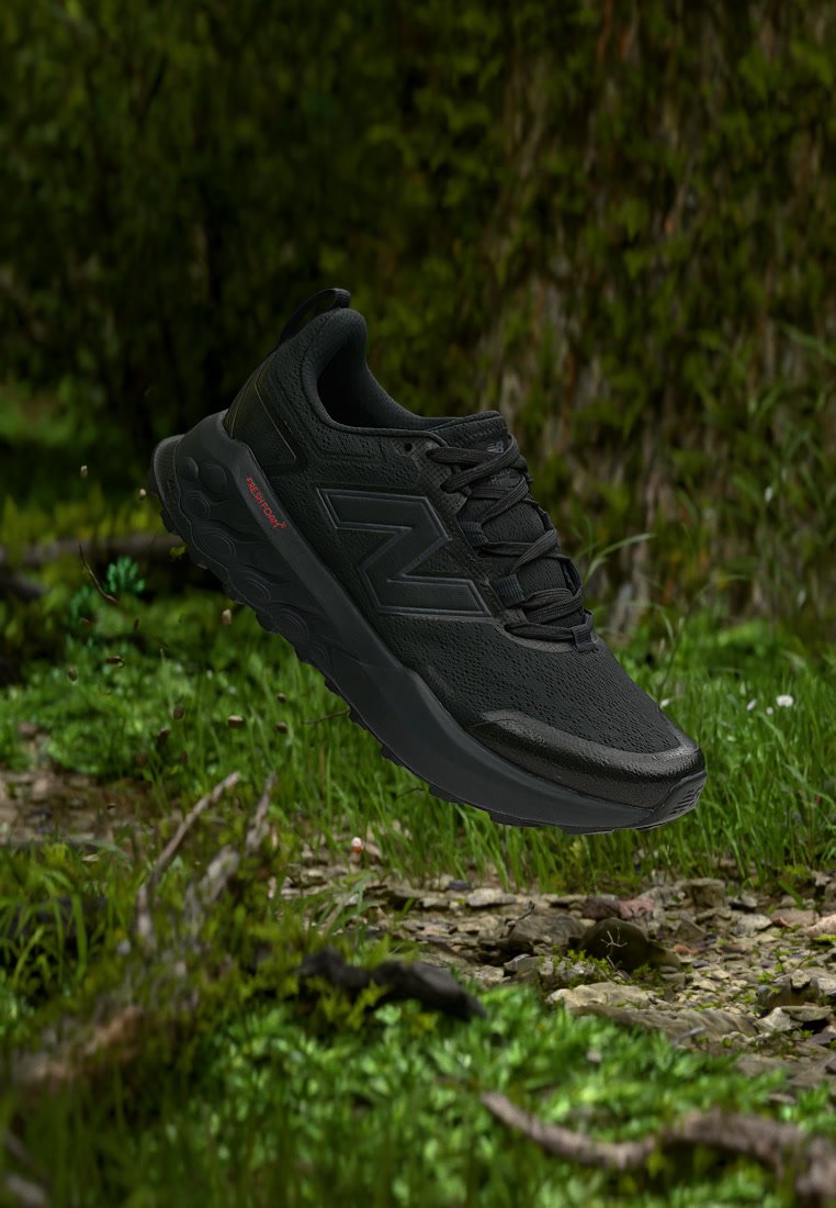 Zapatilla de correr negra con suela texturizada y cordones, flotando sobre el suelo verde del bosque con hierba y pequeñas plantas.