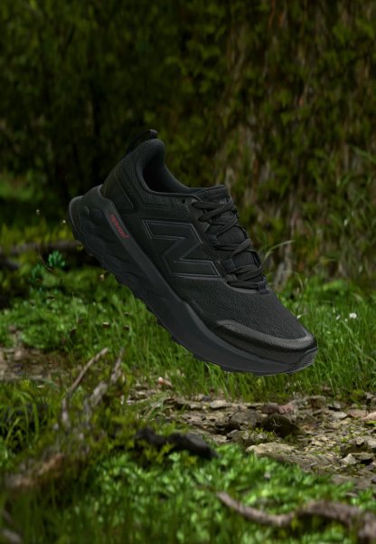Zapatilla de correr negra con suela texturizada y cordones, flotando sobre el suelo verde del bosque con hierba y pequeñas plantas.