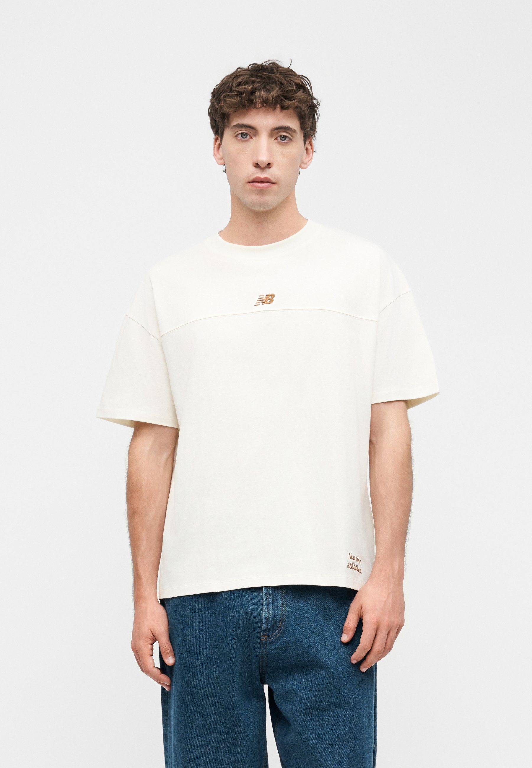 Joven con cabello corto y rizado lleva una camiseta blanca de New Balance y vaqueros azules, de pie frente a un fondo gris liso.