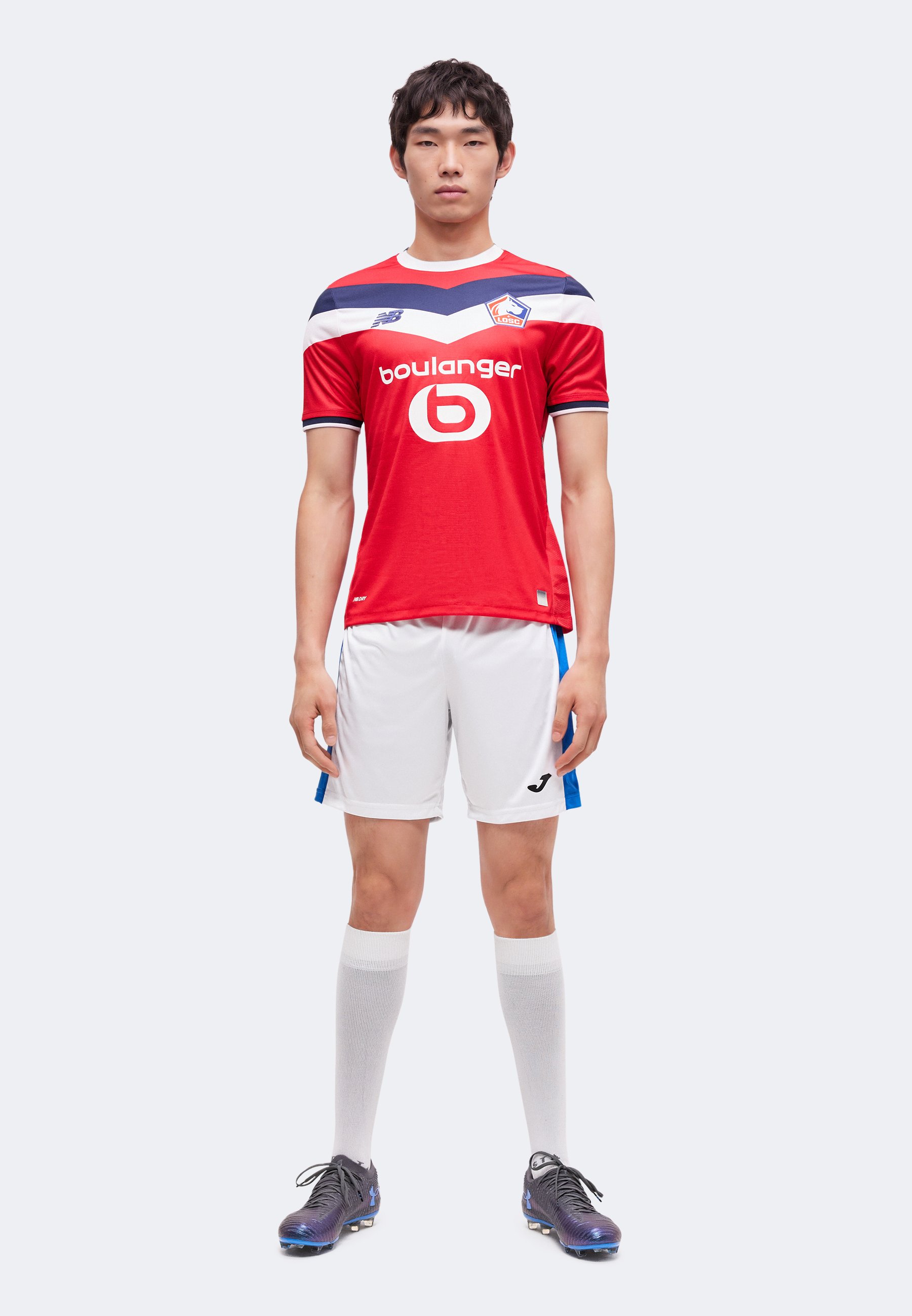 Hombre de pie mirando hacia adelante, vistiendo una camiseta roja del Lille, pantalones cortos blancos con rayas azules, calcetines blancos hasta la rodilla y botas de fútbol negras.