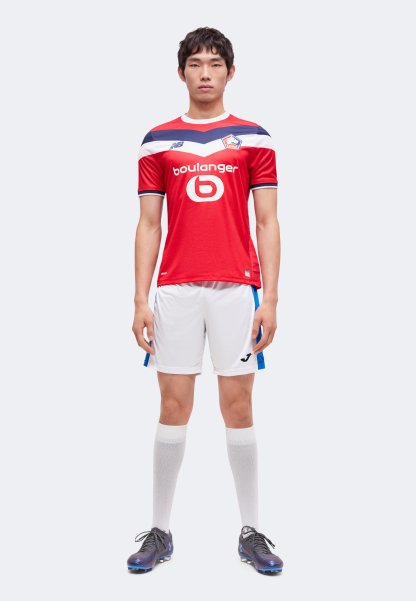 Hombre de pie mirando hacia adelante, vistiendo una camiseta roja del Lille, pantalones cortos blancos con rayas azules, calcetines blancos hasta la rodilla y botas de fútbol negras.