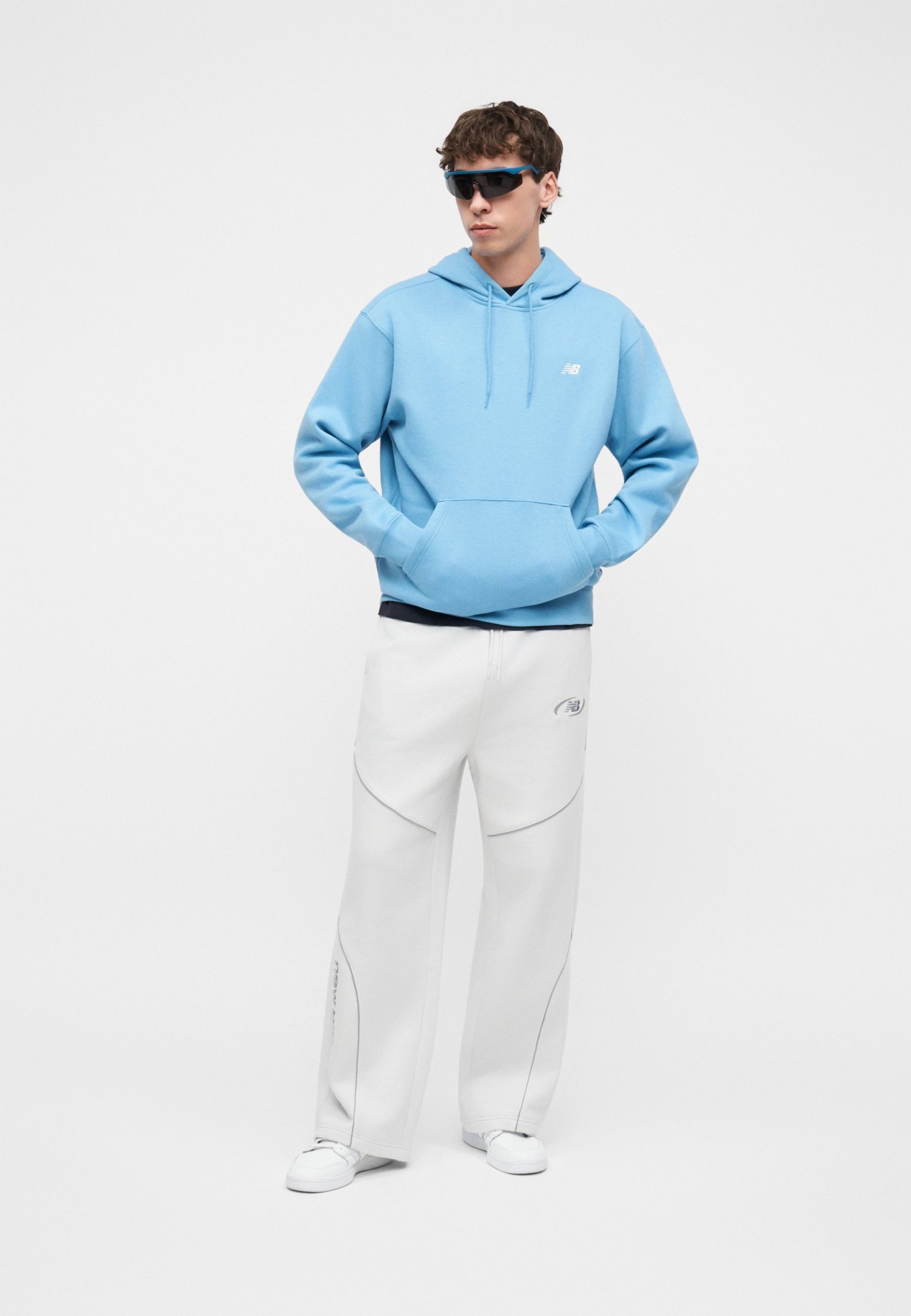 Joven con sudadera azul con capucha, pantalones blancos de pierna ancha, zapatillas blancas y gafas de sol deportivas negras, de pie con las manos en los bolsillos.