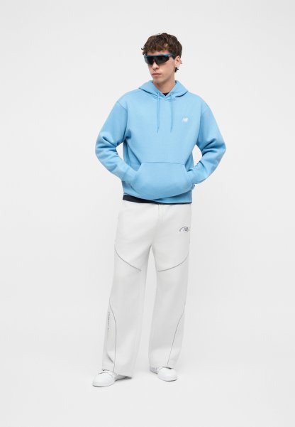 Joven con sudadera azul con capucha, pantalones blancos de pierna ancha, zapatillas blancas y gafas de sol deportivas negras, de pie con las manos en los bolsillos.