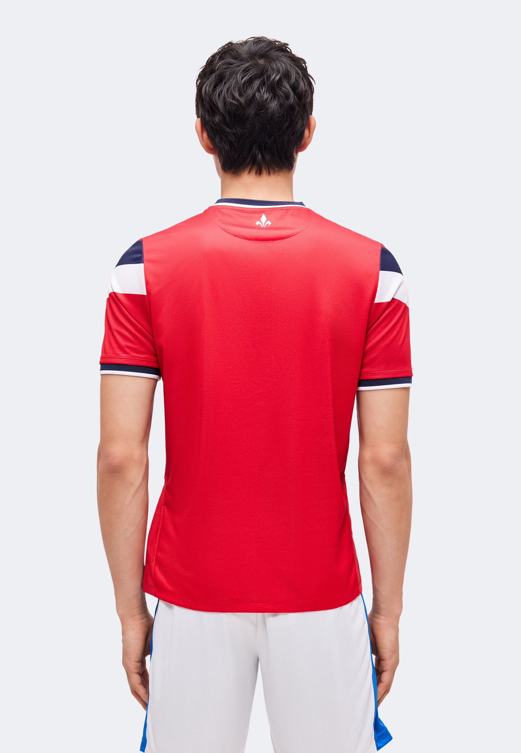 Hombre de pie con la espalda hacia la cámara, vestido con una camiseta deportiva roja con detalles en azul marino y blanco, y pantalones cortos blancos.