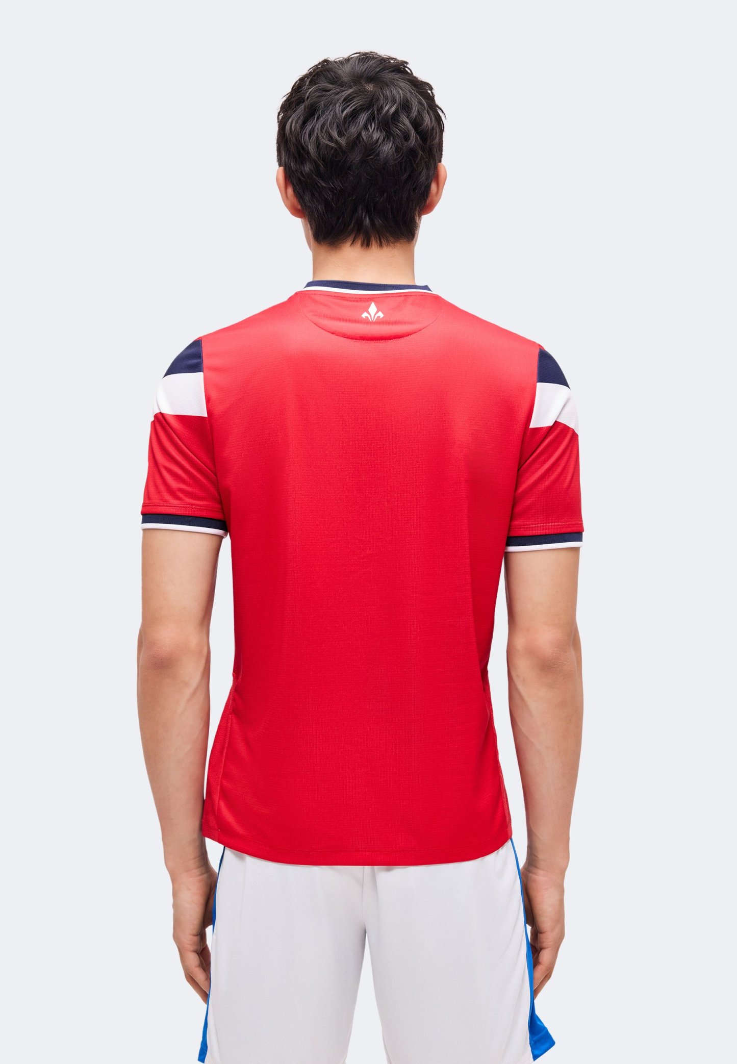 Hombre de pie con la espalda hacia la cámara, vestido con una camiseta deportiva roja con detalles en azul marino y blanco, y pantalones cortos blancos.