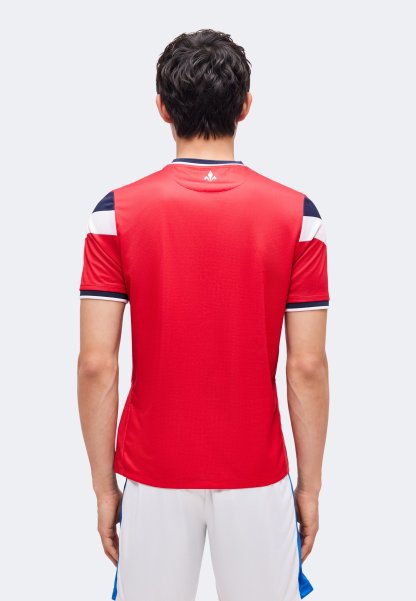 Hombre de pie con la espalda hacia la cámara, vestido con una camiseta deportiva roja con detalles en azul marino y blanco, y pantalones cortos blancos.