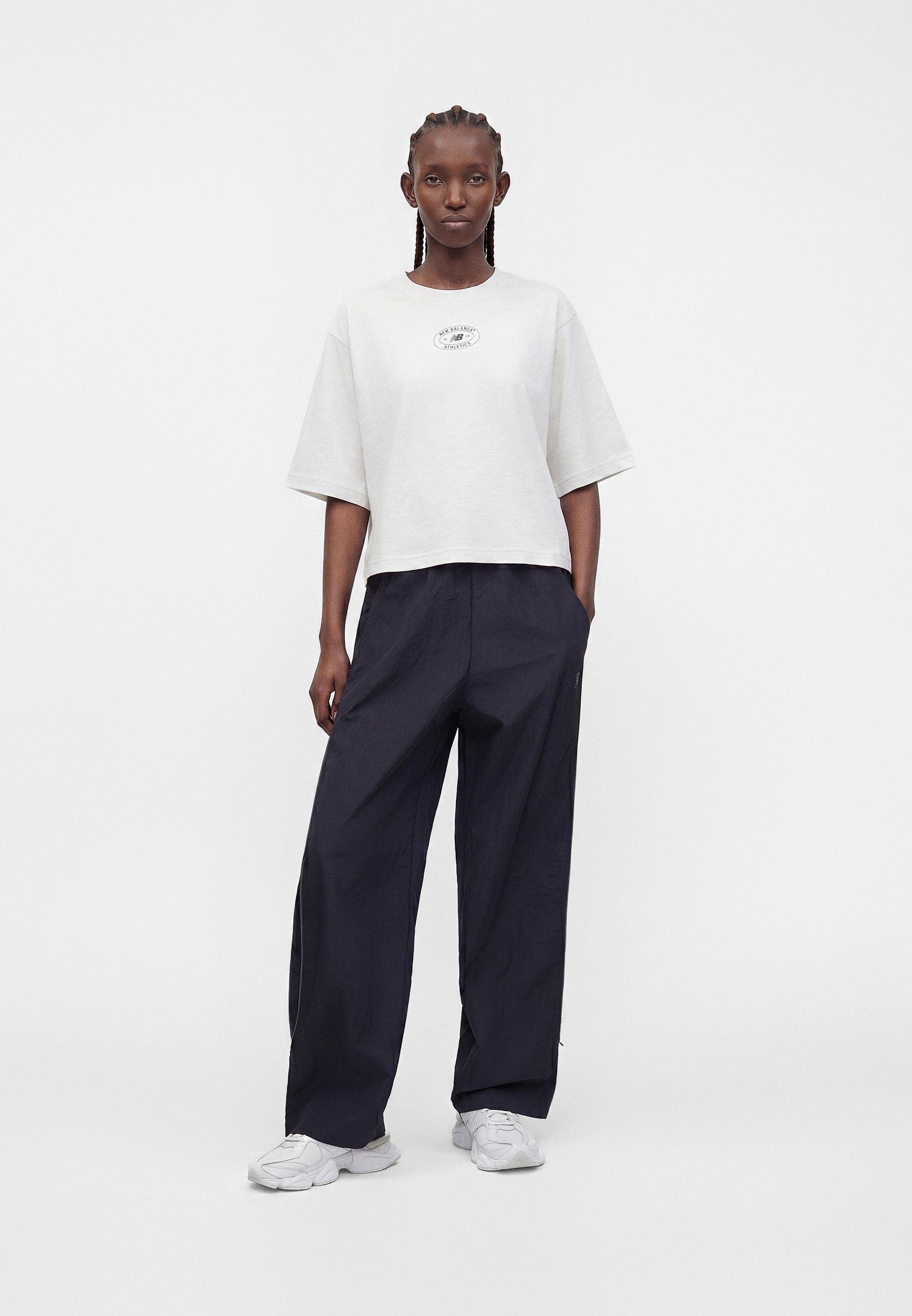 Mujer de pie, mirando hacia adelante, con una camiseta blanca oversize, pantalones negros anchos y zapatillas blancas sobre un fondo blanco liso.