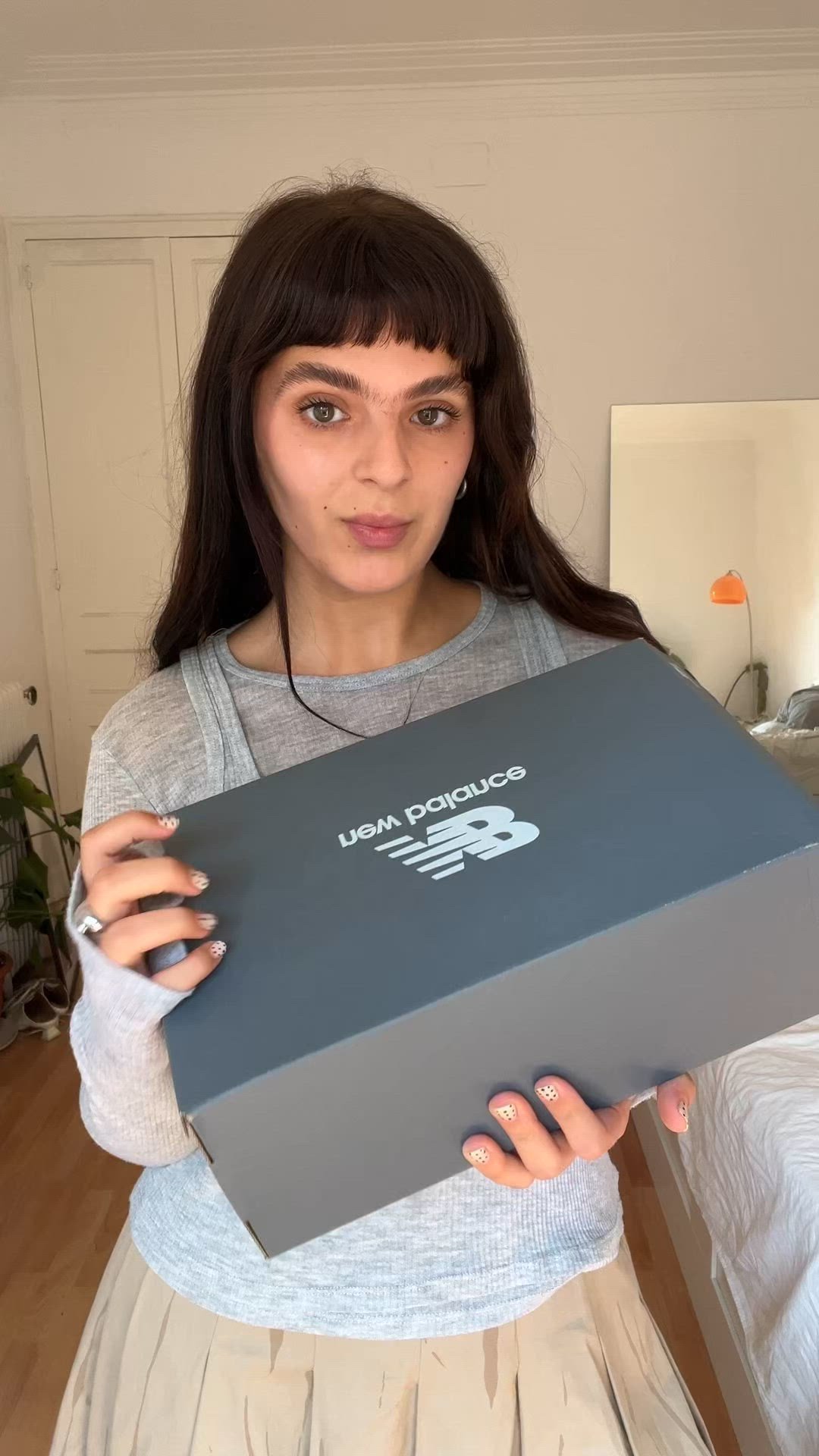 Joven con cabello oscuro sostiene una caja de zapatos New Balance, de pie en un dormitorio con paredes de color claro y suelo de madera.