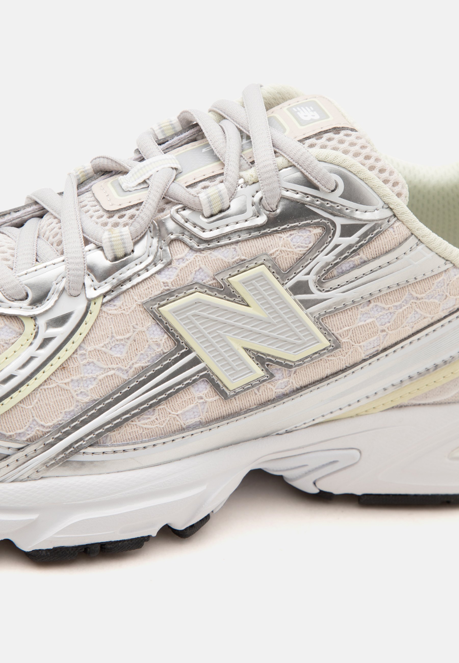 Zapatilla New Balance beige y plateada con detalles de malla y cordones, cordones atados y suela blanca gruesa sobre un fondo liso.