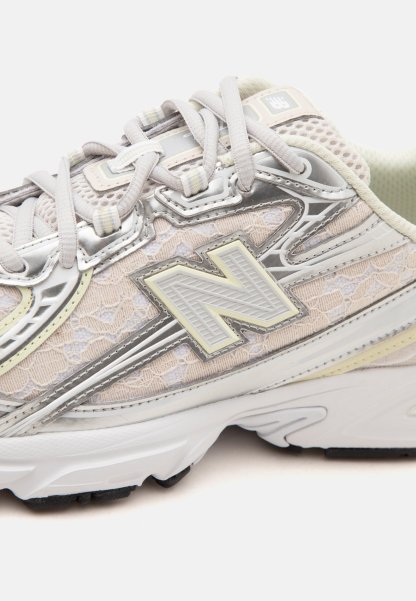 Zapatilla New Balance beige y plateada con detalles de malla y cordones, cordones atados y suela blanca gruesa sobre un fondo liso.