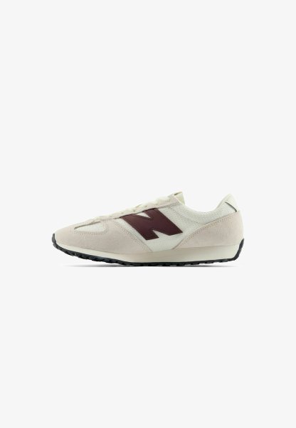Zapatilla blanca y beige con parte superior de ante y malla, logo lateral marrón oscuro, cordones blancos y suela de goma negra mirando hacia la derecha sobre fondo blanco.