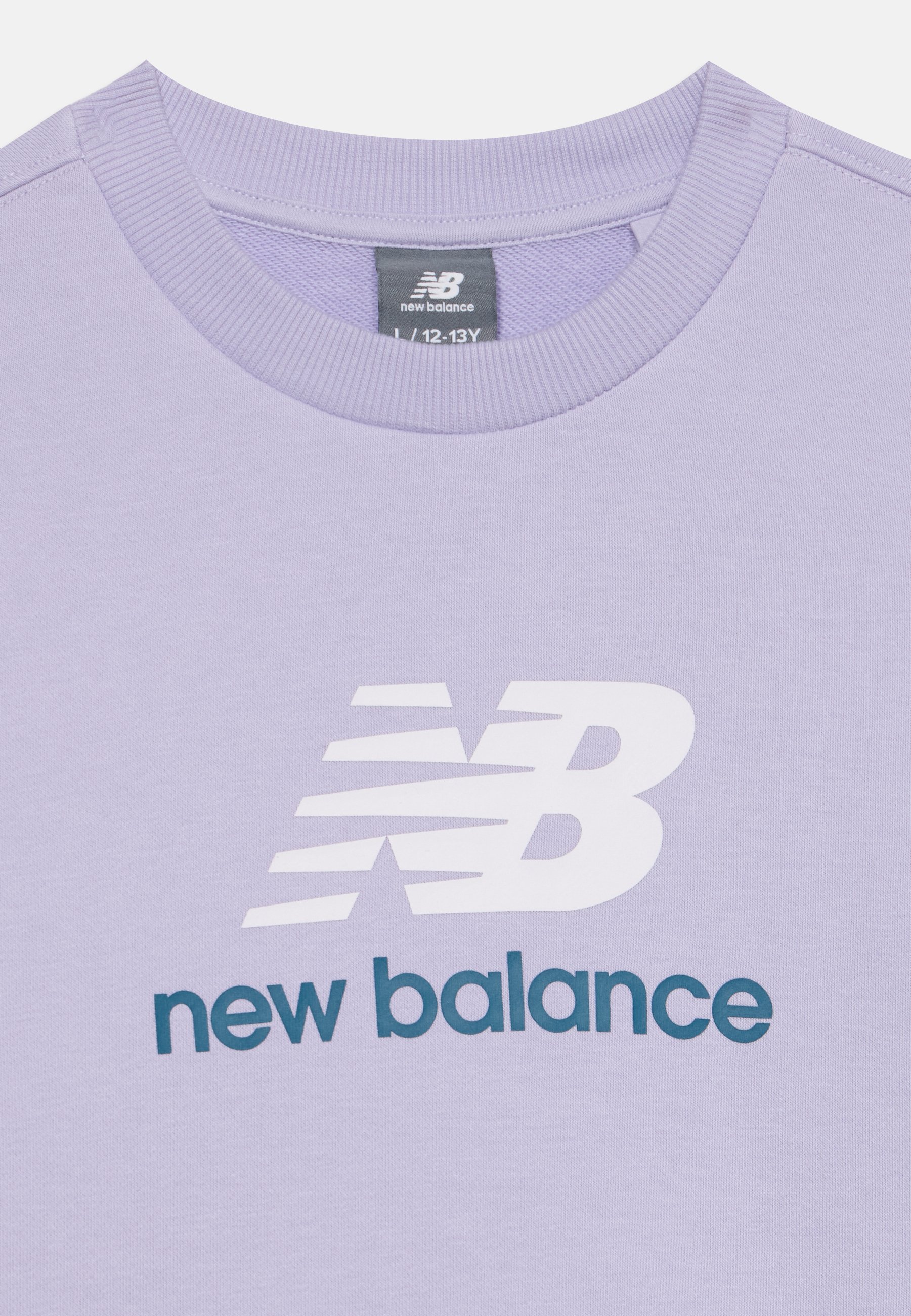 Camiseta de cuello redondo lila con logo blanco de New Balance y texto en verde azulado oscuro "new balance" en la parte delantera, etiqueta de talla 12-13 años visible dentro del cuello.