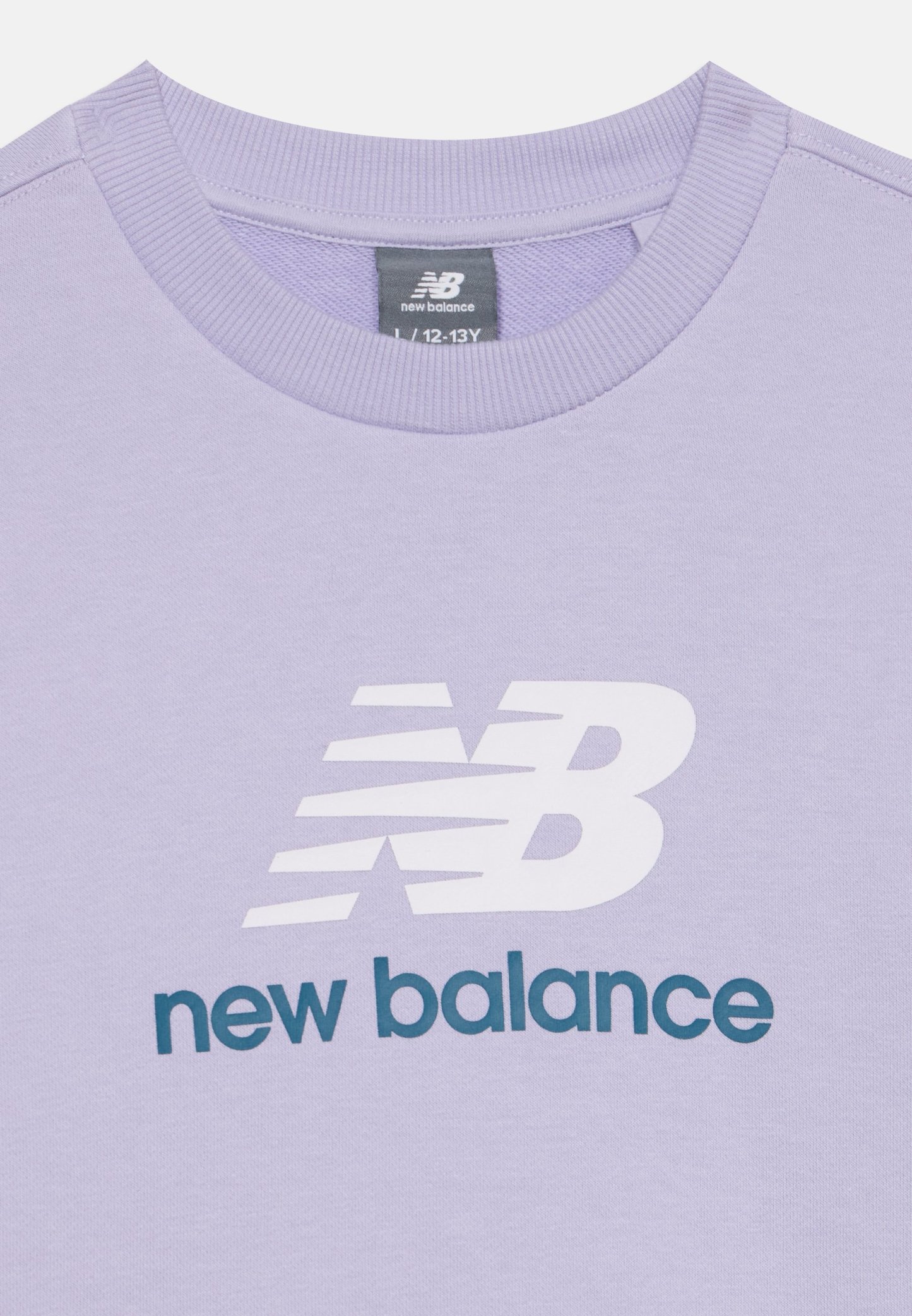 Camiseta de cuello redondo lila con logo blanco de New Balance y texto en verde azulado oscuro "new balance" en la parte delantera, etiqueta de talla 12-13 años visible dentro del cuello.