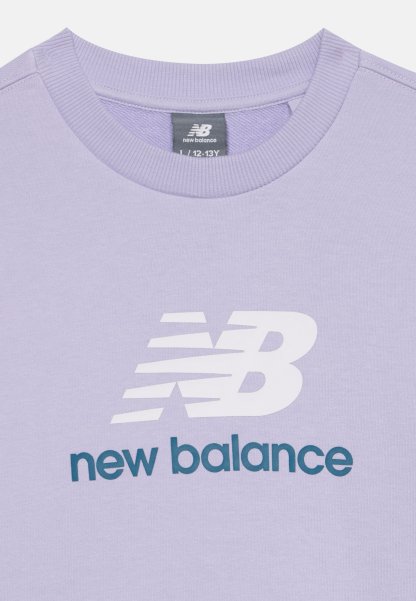 Camiseta de cuello redondo lila con logo blanco de New Balance y texto en verde azulado oscuro "new balance" en la parte delantera, etiqueta de talla 12-13 años visible dentro del cuello.