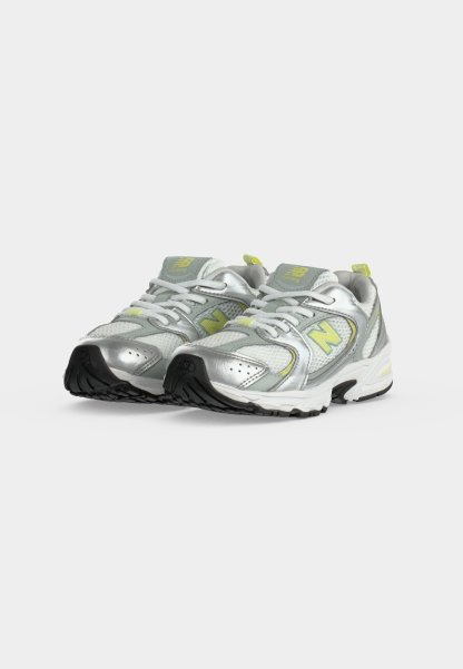 Par de zapatillas de correr plateadas y grises con detalles amarillos y suelas negras, colocadas lado a lado sobre un fondo blanco.