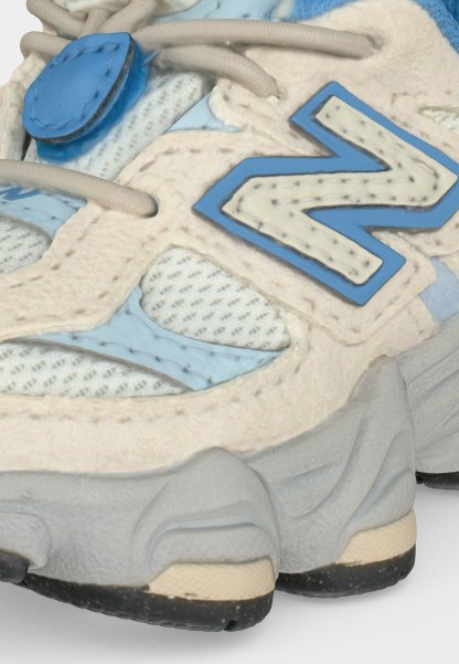 Primer plano de una zapatilla deportiva azul y beige con tejido de malla, superposiciones de ante, suela gruesa y texturizada, y un logo "N" cosido en el lateral.
