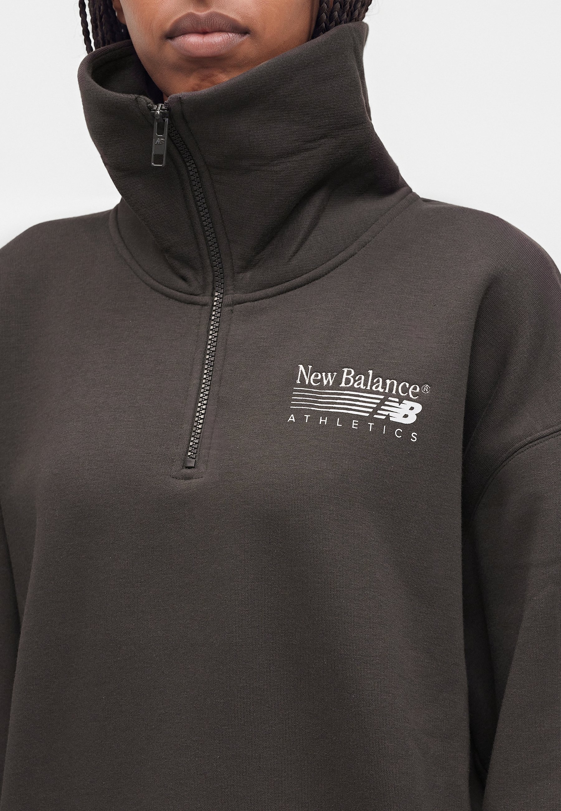 Persona que lleva un jersey oscuro de New Balance Athletics con cremallera de un cuarto y cuello alto, enfocándose en el torso superior y el logo.