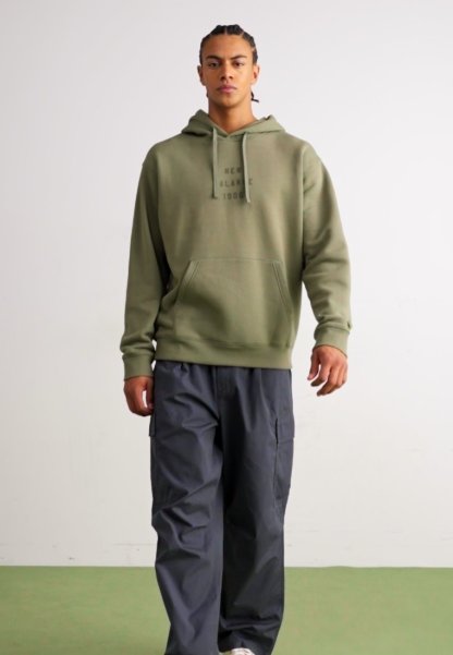 Sudadera verde oliva con bolsillo frontal y capucha con cordón, combinada con pantalones cargo gris oscuro que tienen bolsillos laterales.