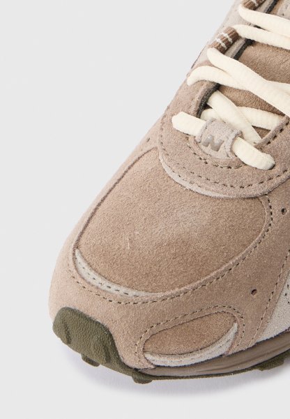 Acercamiento a la puntera de una zapatilla de ante beige con cordones crema, costuras detalladas y un borde de suela de goma verde oliva texturizada.