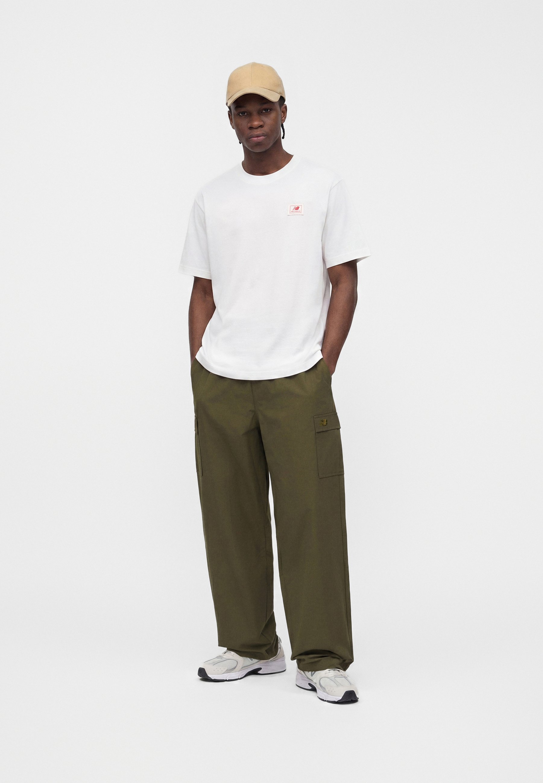 Hombre con gorra beige, camiseta blanca de manga corta, pantalones cargo verde oliva y zapatillas blancas, de pie con las manos en los bolsillos.