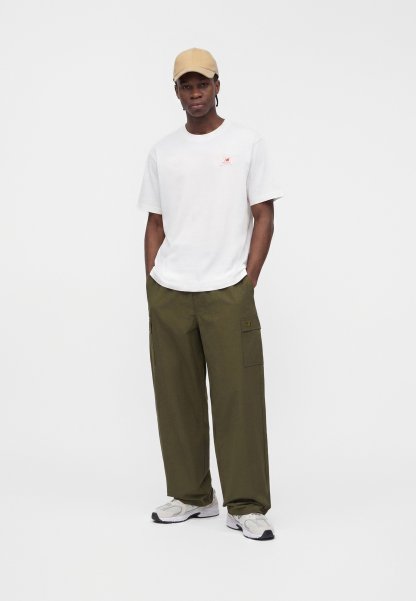 Hombre con gorra beige, camiseta blanca de manga corta, pantalones cargo verde oliva y zapatillas blancas, de pie con las manos en los bolsillos.