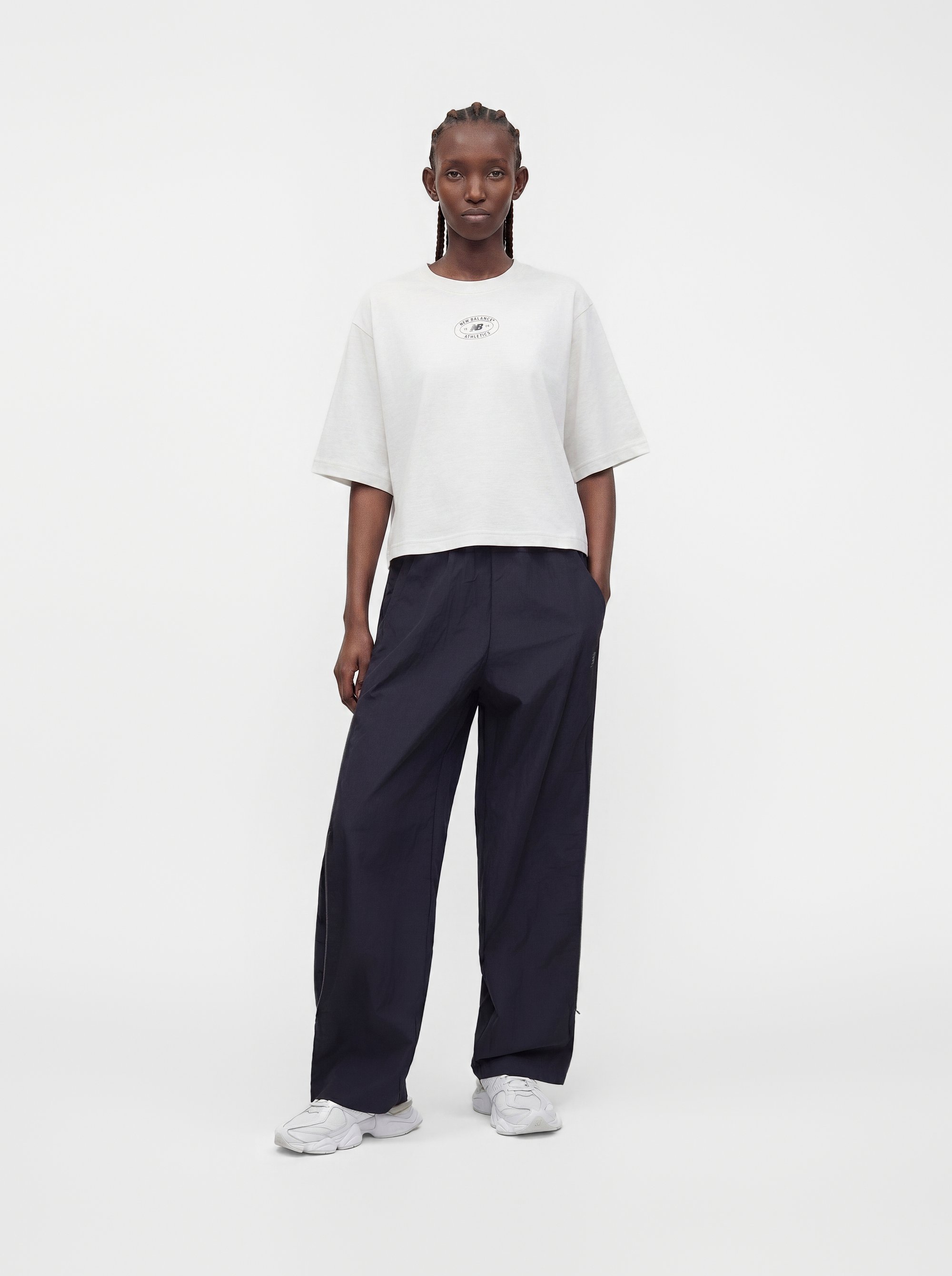 Persona de pie con trenzas, vistiendo una camisa blanca de manga corta oversize, pantalones anchos oscuros y zapatillas blancas sobre un fondo liso.