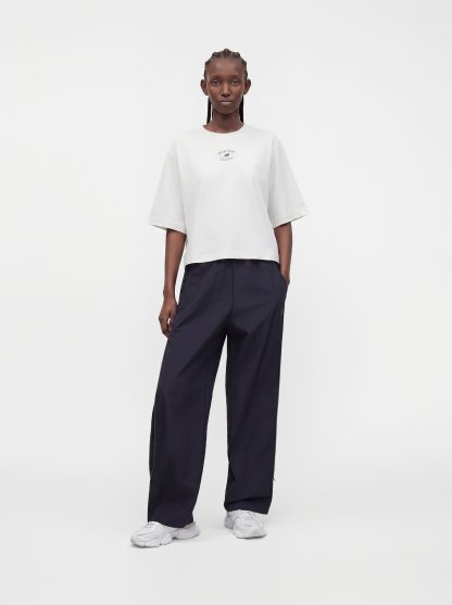 Persona de pie con trenzas, vistiendo una camisa blanca de manga corta oversize, pantalones anchos oscuros y zapatillas blancas sobre un fondo liso.