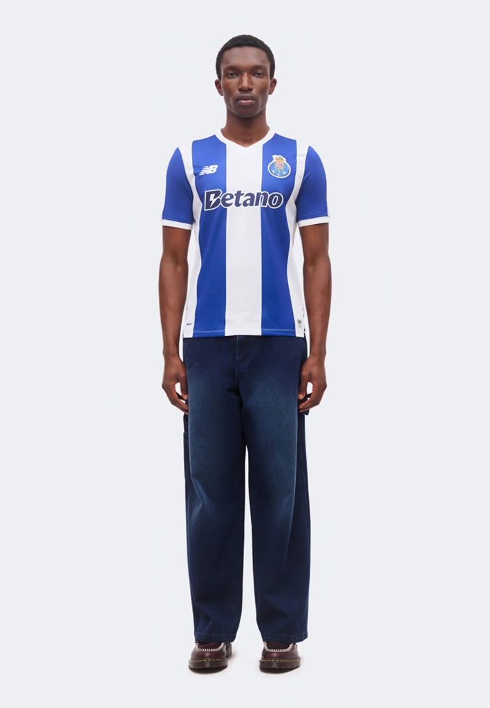 Hombre de pie vistiendo una camiseta de fútbol a rayas verticales azules y blancas con el logo de "Betano", pantalones anchos oscuros y zapatos marrones.