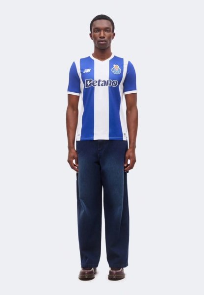 Hombre de pie vistiendo una camiseta de fútbol a rayas verticales azules y blancas con el logo de "Betano", pantalones anchos oscuros y zapatos marrones.