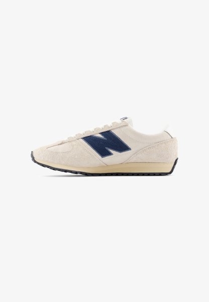 Zapatilla deportiva con parte superior de ante beige, logo azul marino, detalles blancos y suela de goma negra. Cuenta con cordones planos y diseño aerodinámico.
